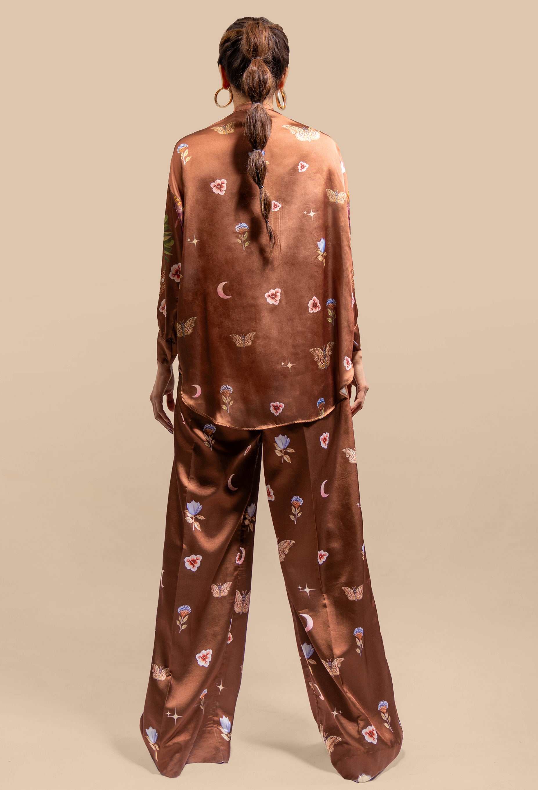 A model wearing the Cosmic Print Silk Co-Ord Set, standing confidently in the blouse and matching trousers. The flowing silk blouse is tucked slightly into the high-waisted trousers, creating a streamlined silhouette. The cosmic print adds a dynamic, artistic flair to the outfit, which the model complements with minimal accessories and soft makeup.